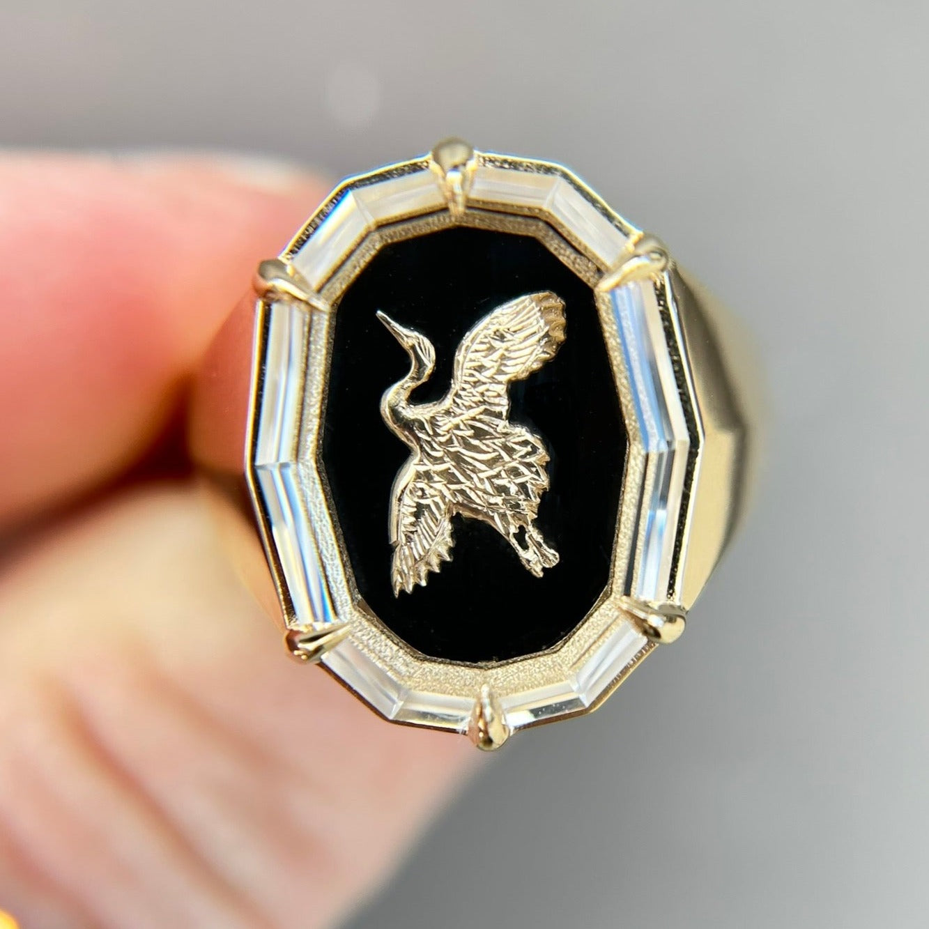 A signet ring with a gold bird emblem on a black onyx background, held between fingers. The gold Japanese crane contrasts boldly against the gold - creating a breathtaking and unique custom ring.