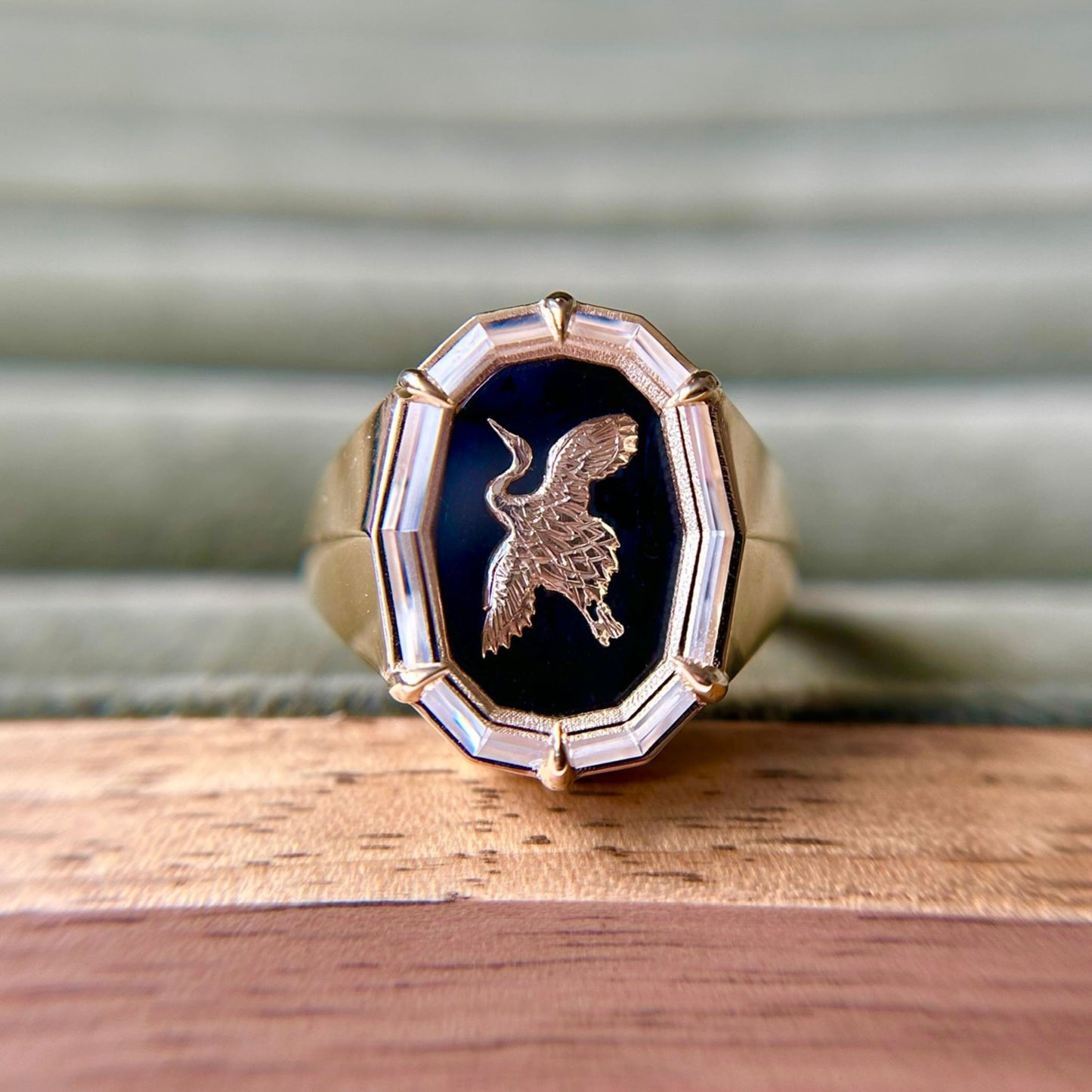 Gold ring with a black onyx inlay featuring an engraved bird, framed by a faceted border. It's placed on a wooden surface with a green, textured background.