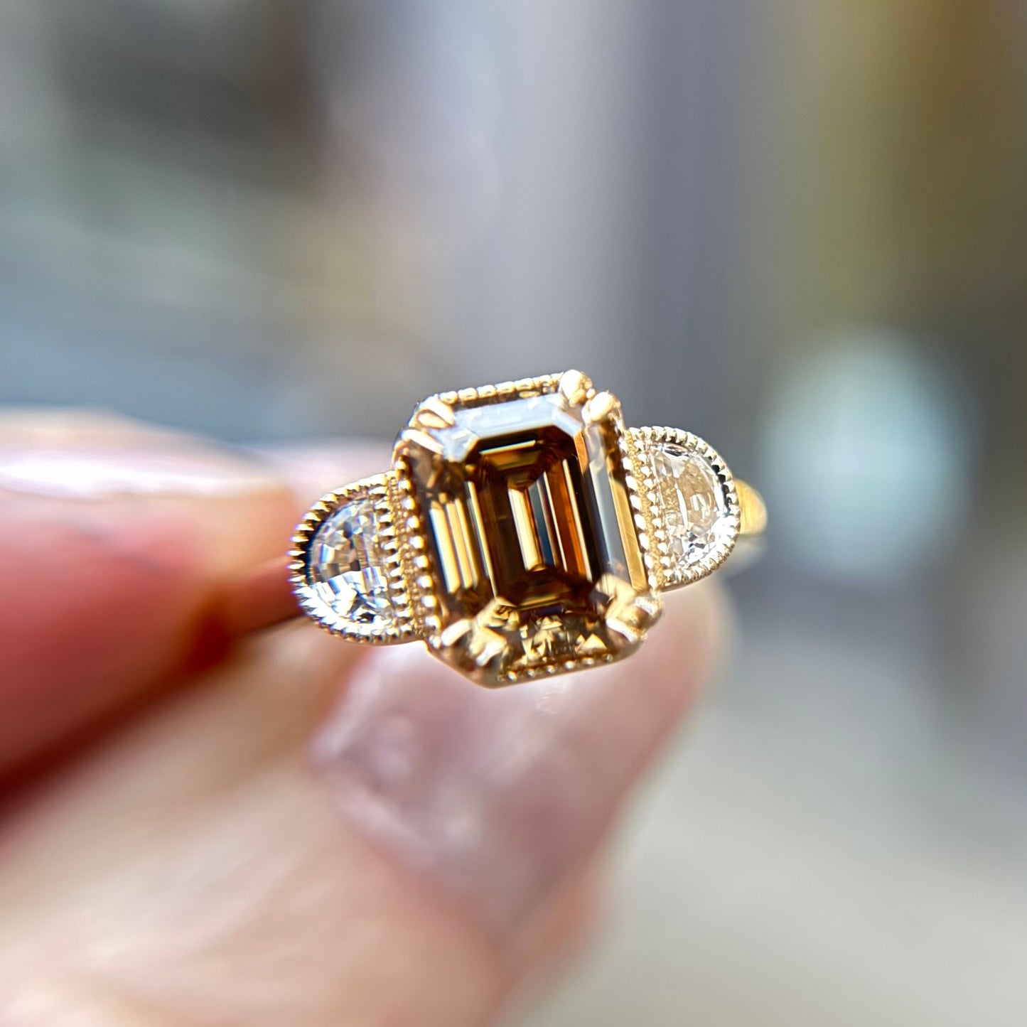A hand holds a gold ring with an emerald-cut, champagne diamond centered between two smaller round diamonds. The background is softly blurred.