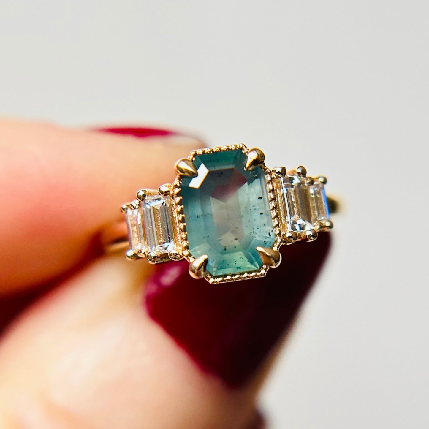 A gold ring with a large, rectangular light teal gemstone at the center, flanked by two smaller rectangular clear stones. A hand with red nails holds it.