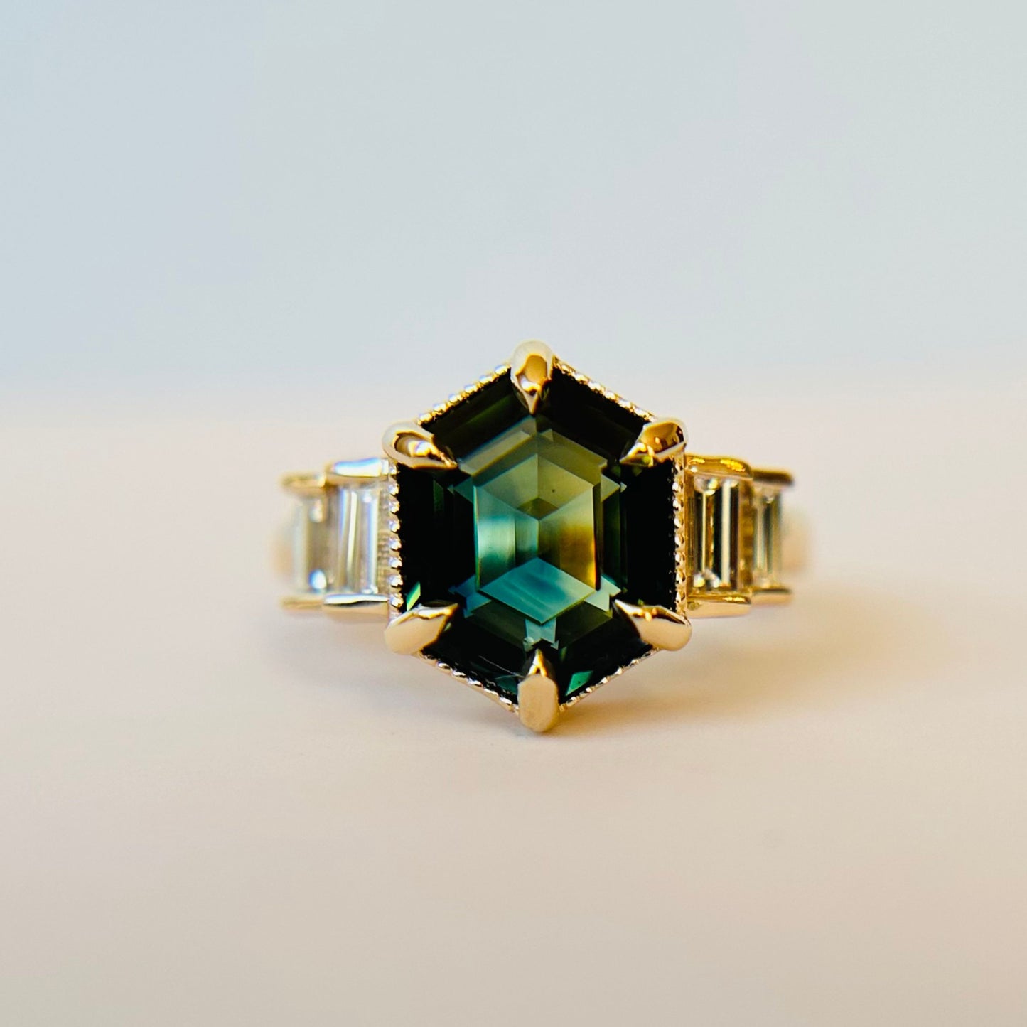 A hexagonal green sapphire set in a gold engagement ring with rectangular diamonds on the sides. The deep green hues exude elegance and luxury against a plain background.