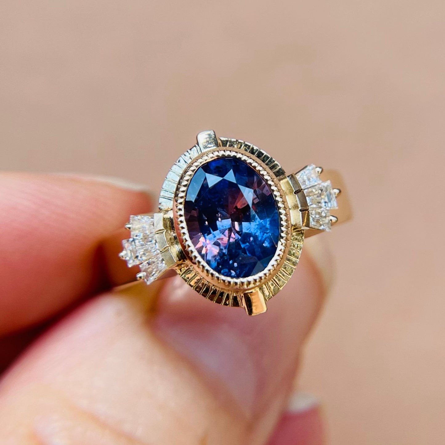 A hand holds an elegant ring featuring an oval, dark blue almost purple sapphire set in gold, flanked by two clusters of small, sparkling diamonds, against a soft beige background.