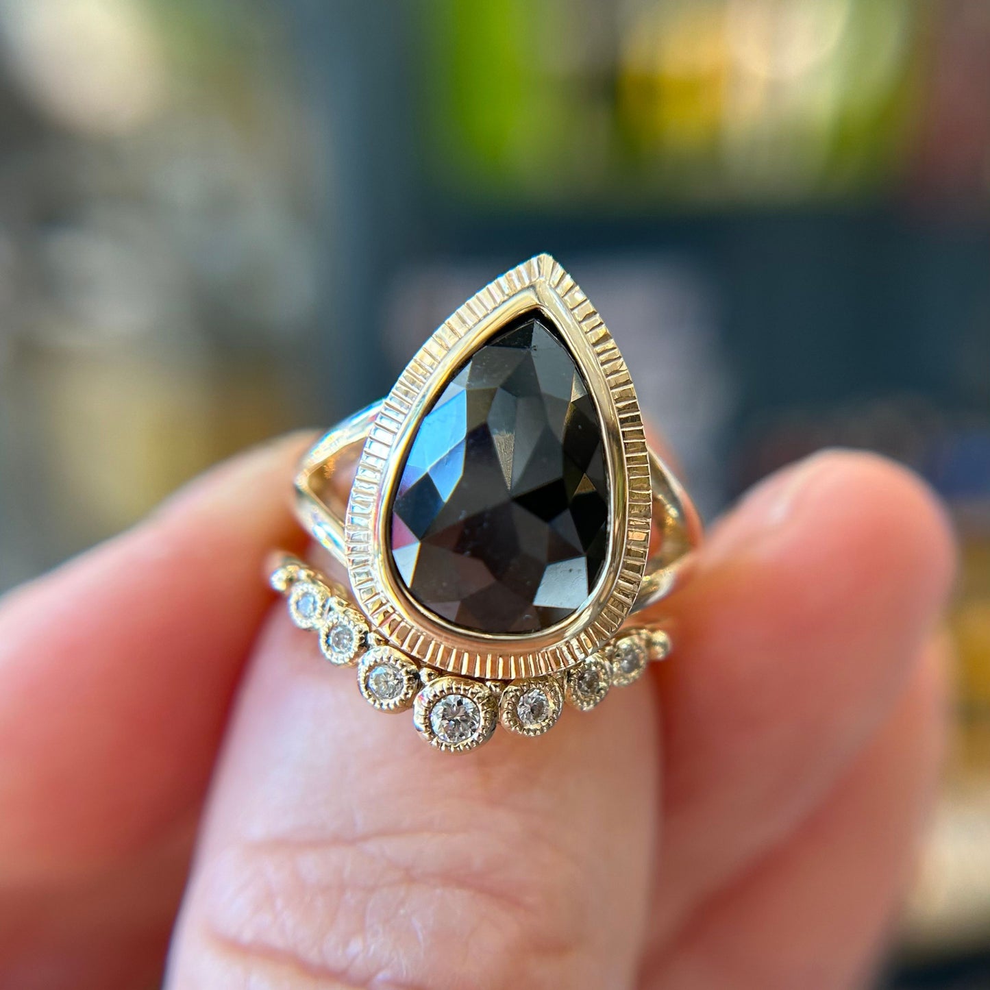 A hand holds a gold ring with a large, pear shaped black diamond, stacked together with a diamond contoured wedding band, against a blurred background.