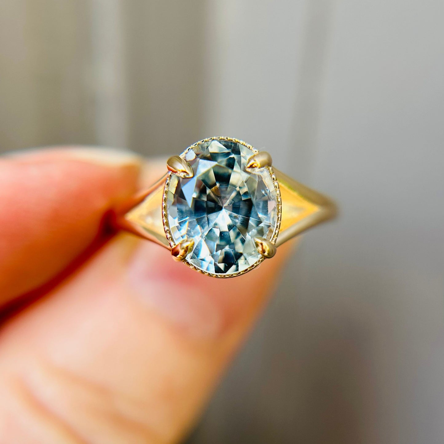 Close-up of a gold ring with an oval-cut blue sapphire held between fingers. The facets reflect light, creating a sparkling and elegant appearance.