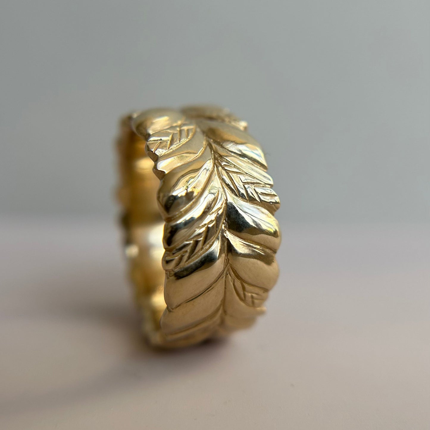 A 14k gold ring with intricate leaf patterns stands on its edge against a soft gray background. The design exudes elegance and a natural charm. Perfect for a wedding ring or other stacking band.