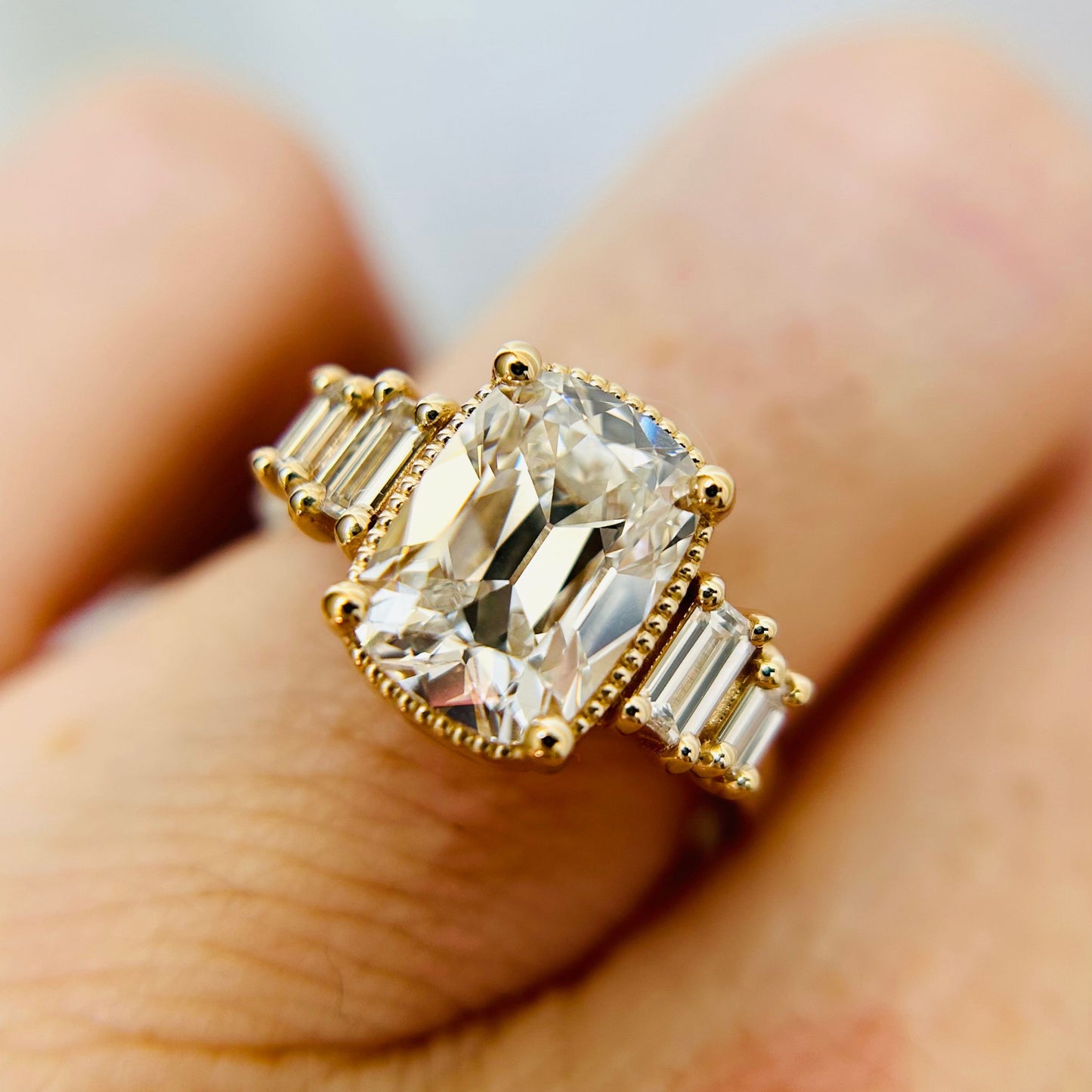 Close-up of a gold engagement ring with a large central oval diamond and baguette diamond accents, worn on a finger. The setting is inspired by art-deco fashion and is elegant and sophisticated.