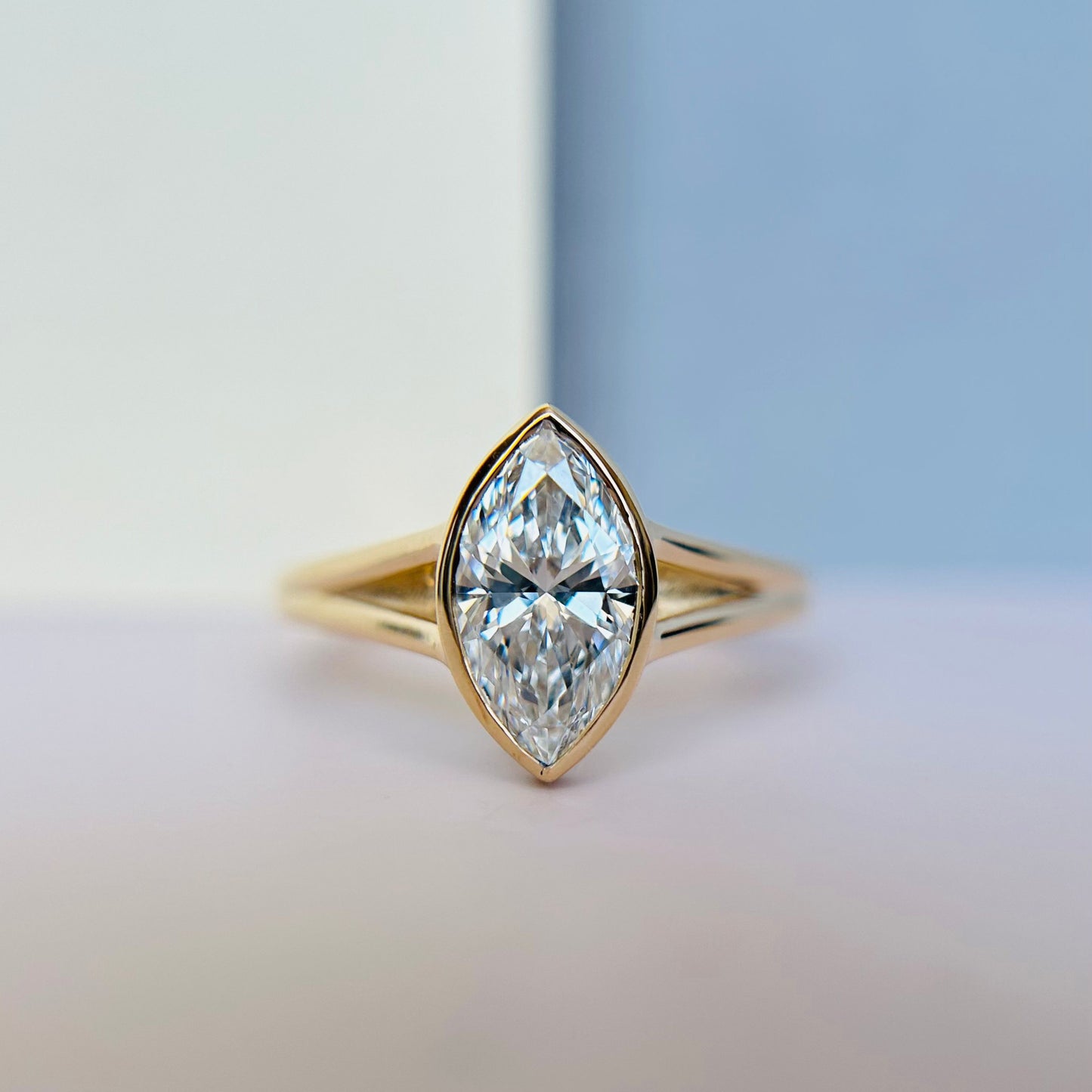 A marquise-cut diamond ring with a gold split-shank band is centered against a gradient background of white and blue, conveying elegance and luxury.