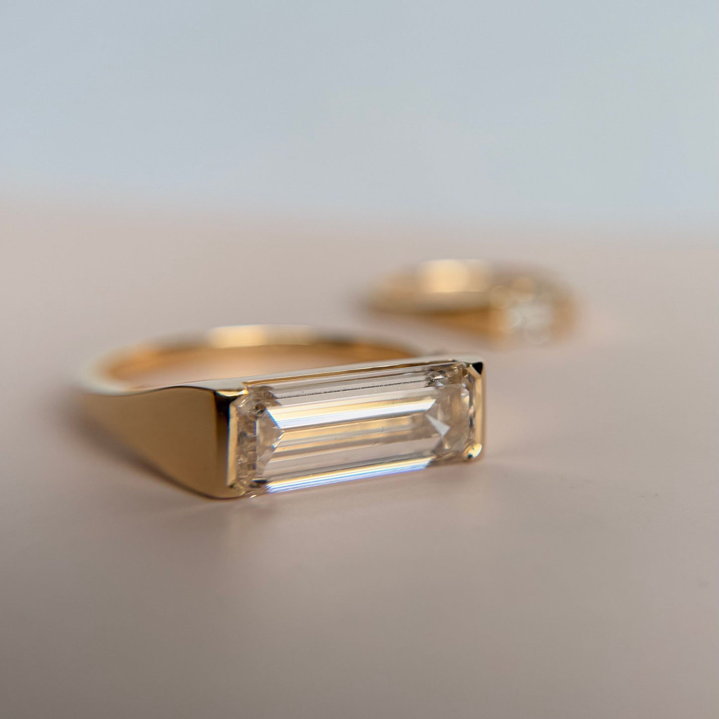 Gold ring with an emerald-cut diamond on a soft pink surface, casting a subtle shadow. Another blurred ring is visible in the background.