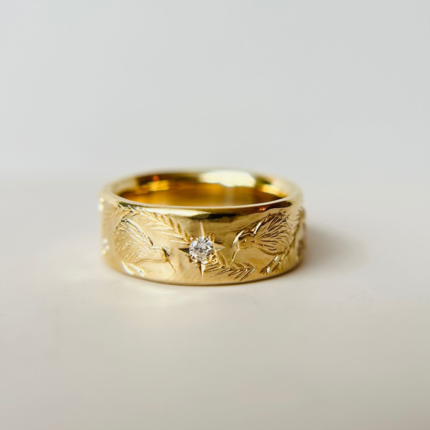 A wide gold ring with hand carved pine branches, birds and a small centered diamond. The ring has a polished finish, conveying elegance and artistic flair.