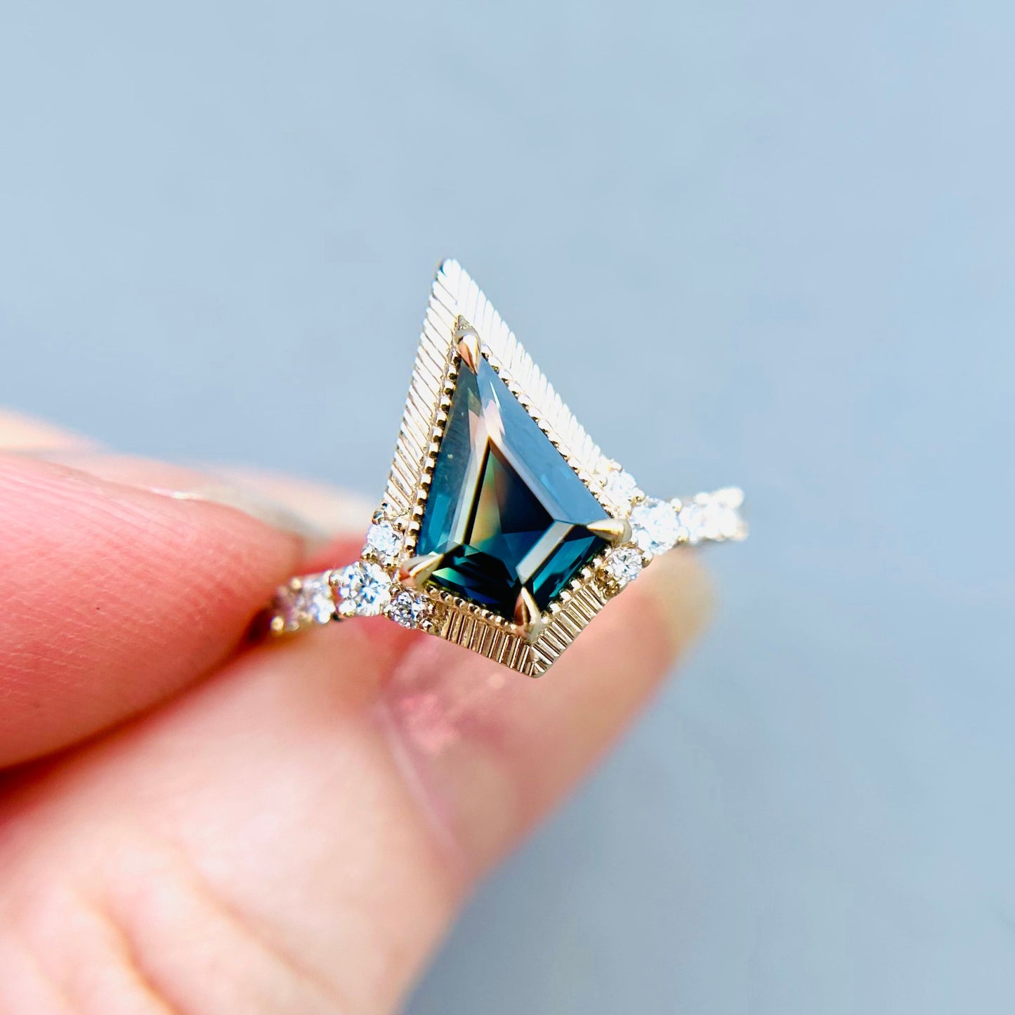 A hand holds an engagement ring with a triangular deep blue-green gemstone, set in gold with a textured frame and a band adorned with small sparkling diamonds. The ring is unique and geometric in style.