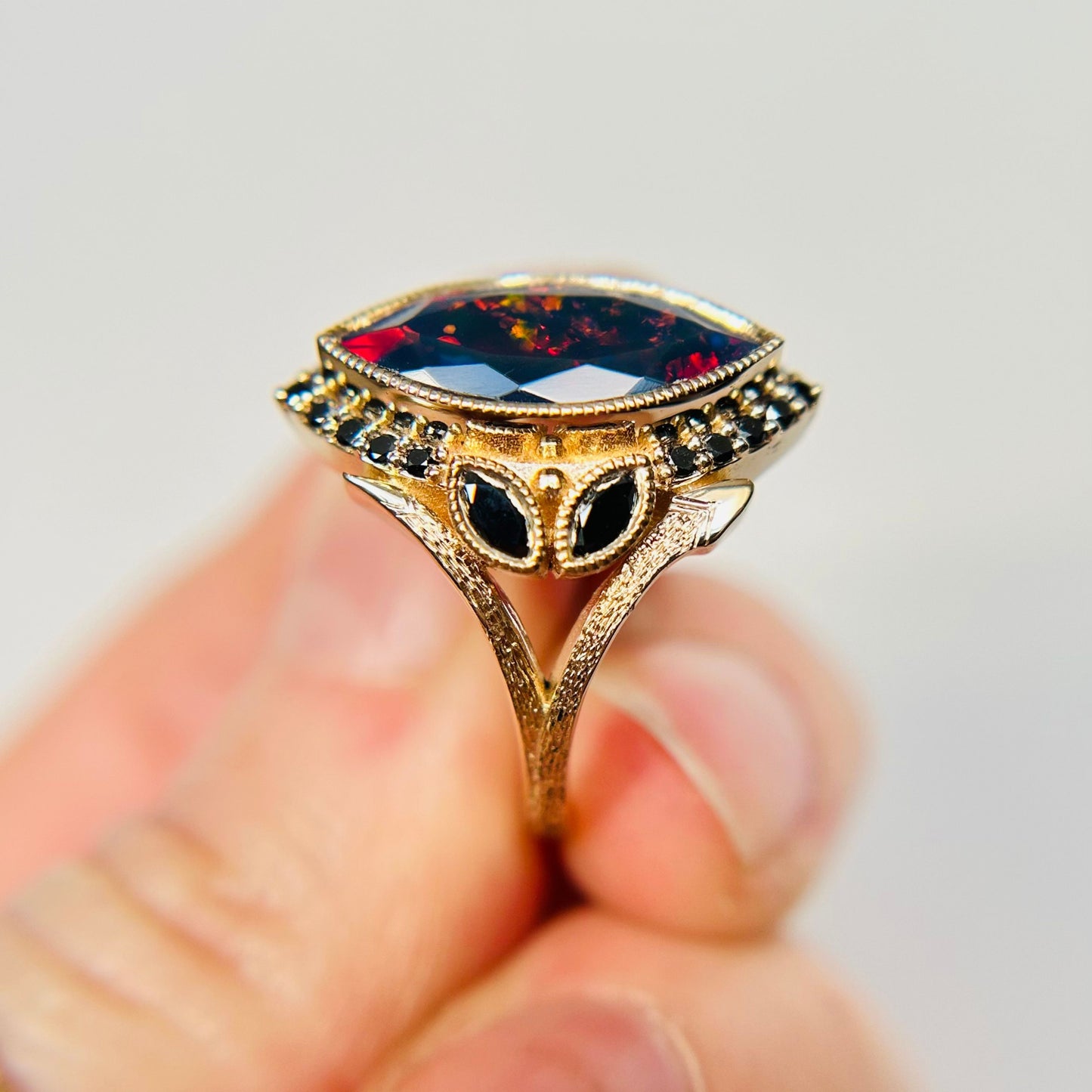Close-up of a hand holding an ornate gold ring with a large, faceted marquise-shaped red opal flanked by a halo of small black diamonds, exuding unique taste, artistry elegance.