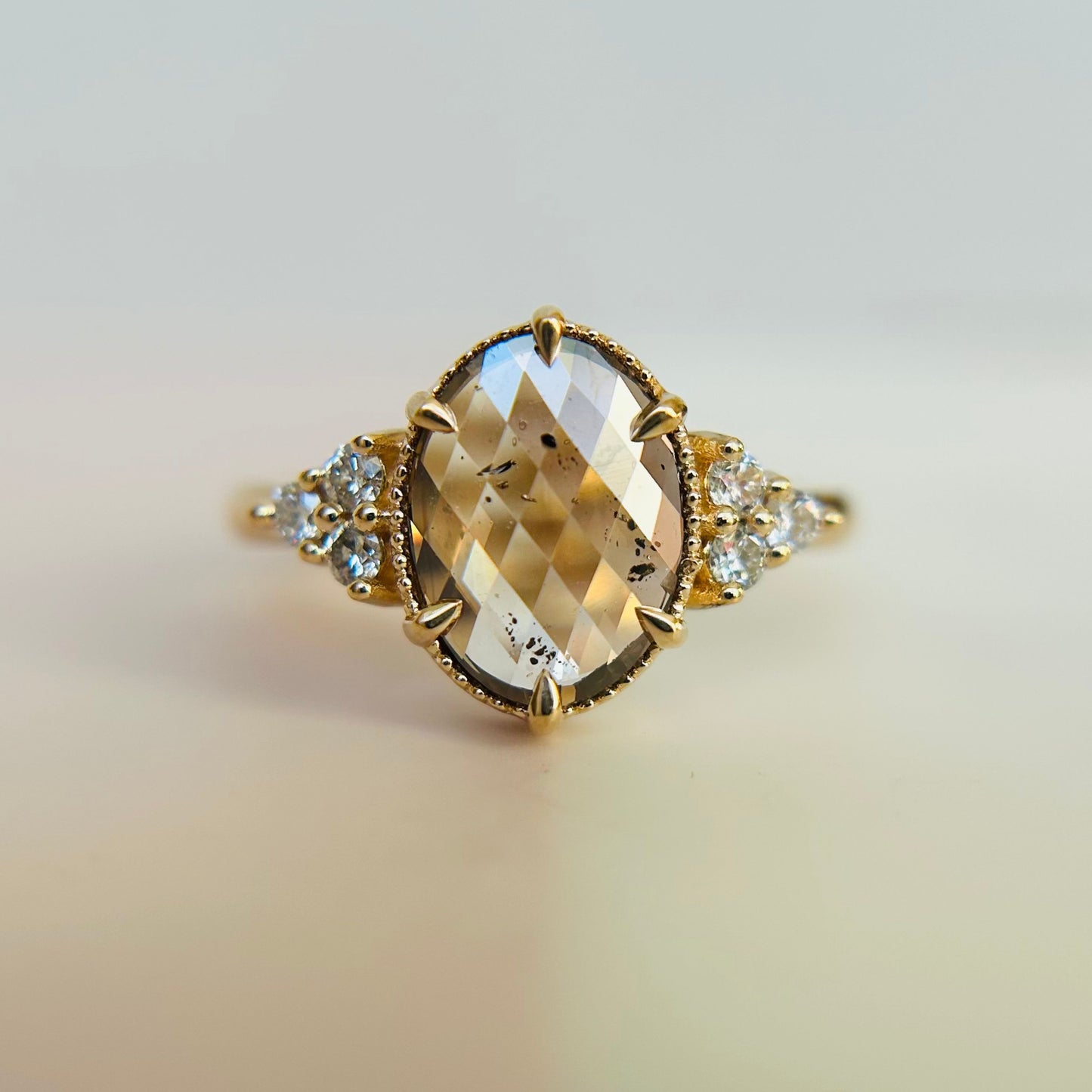 A gold engagement ring features a large, faceted oval diamond with a reflective surface. Small round diamonds flank the centerpiece, creating an elegant, luxurious look.