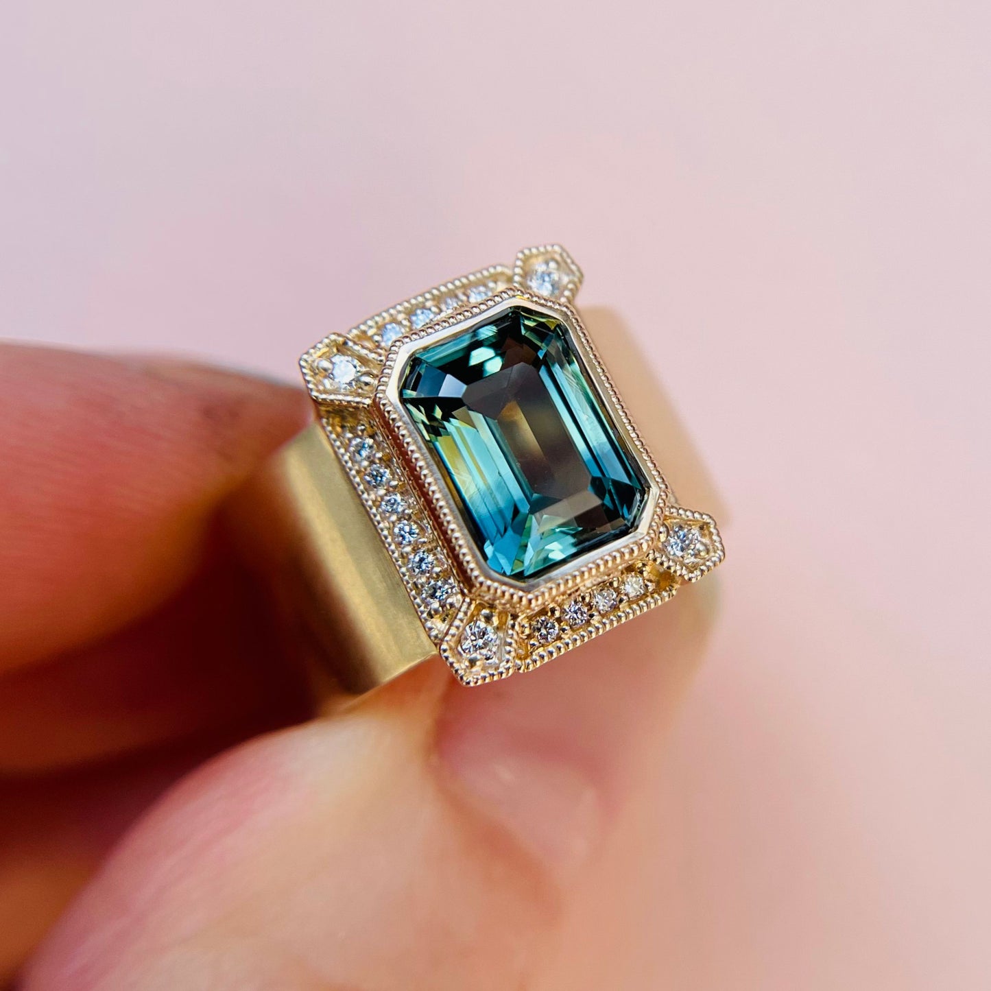 A gold ring with an emerald-cut teal sapphire, surrounded by small sparkling diamonds, held between fingers against a soft pink background. A unique and custom piece.