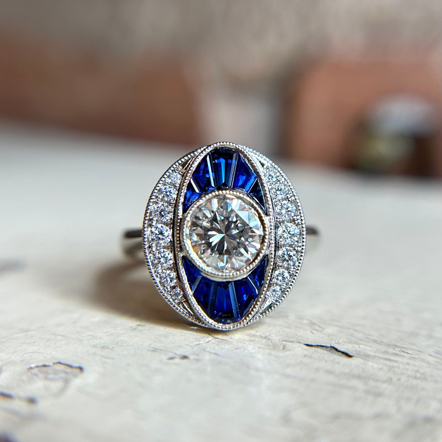 A vintage-style white gold ring featuring a central round diamond, surrounded by deep blue sapphires and smaller diamonds, set on a white surface. Elegant and sparkling.