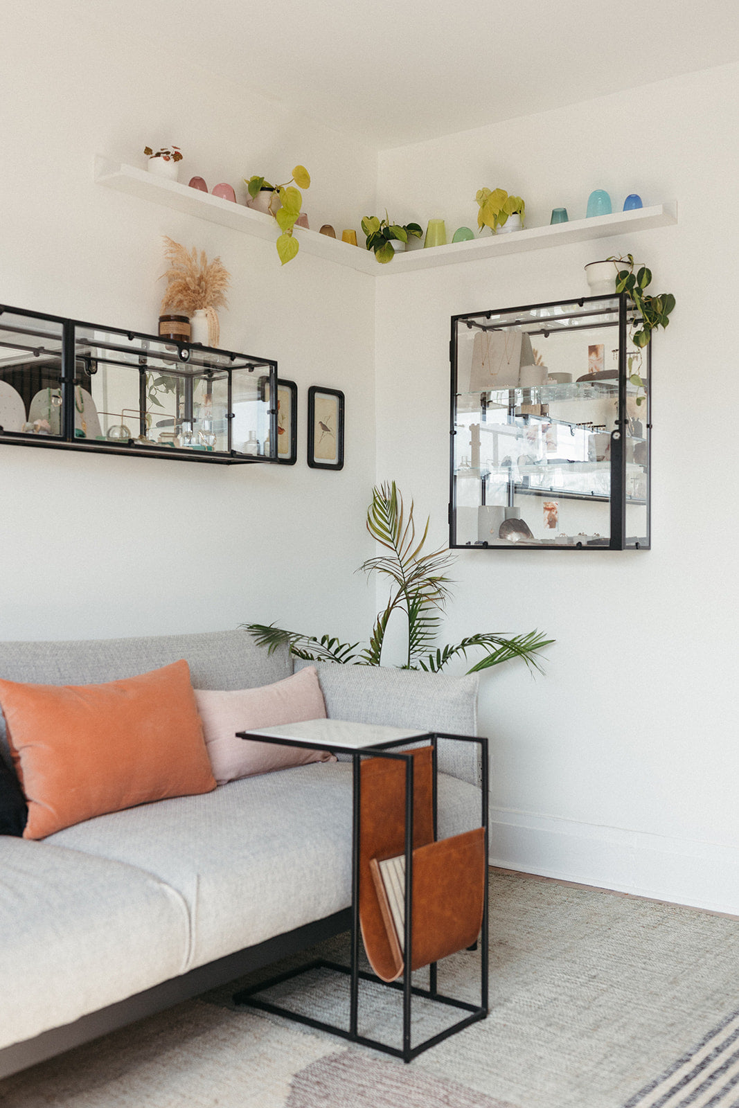 A cozy studio with a gray sofa adorned with orange and pink pillows. A sleek side table holds magazines. Wall shelves display colorful decor and plants.