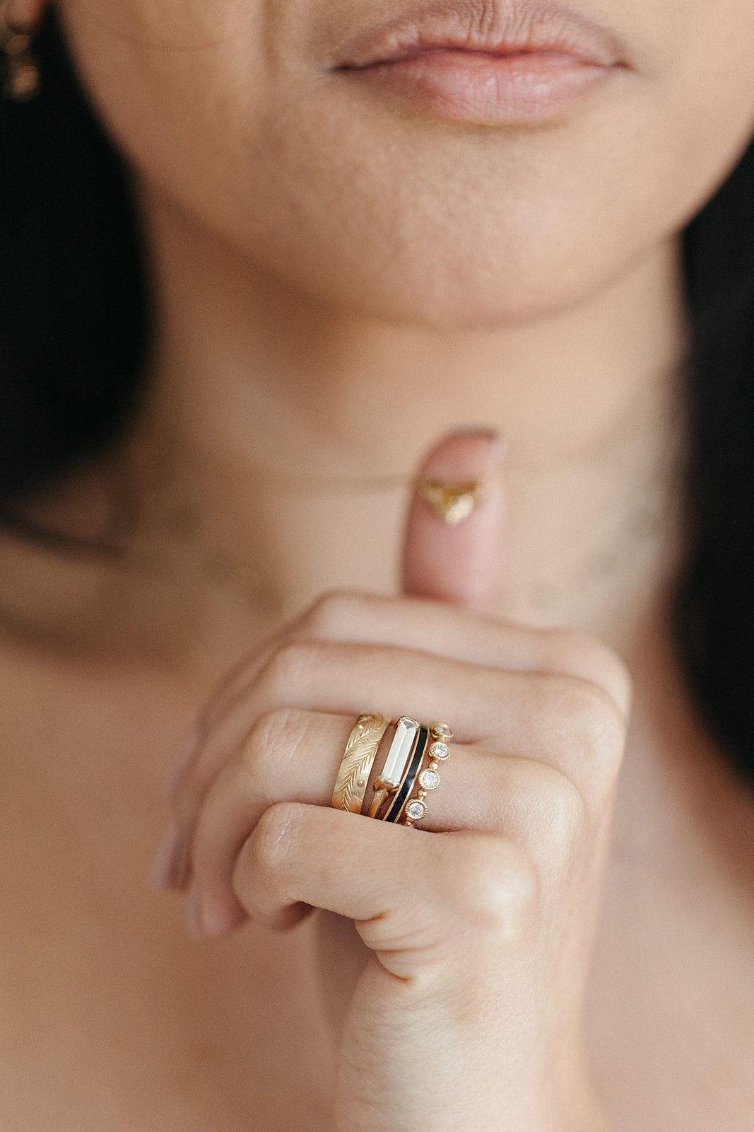 Close-up of a woman's hand touching the necklace around her neck, adorned with gold rings featuring intricate designs and gemstones. The tone is elegant and reflective.