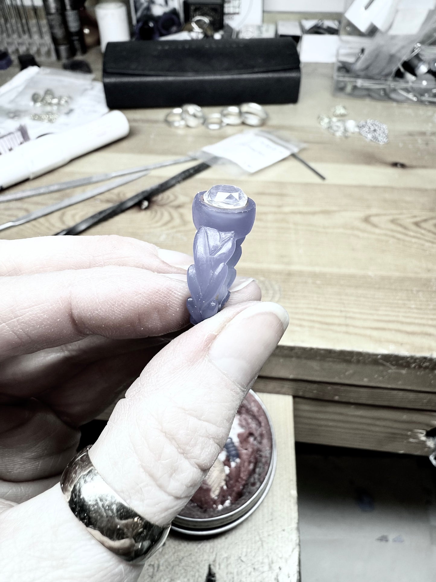 A hand holds up a wax carving of an engagement ring, the wax model holds the centre gemstone in shape and is ready to be sent to be cast in metal. The background shows a jewellery workbench with tools and other jewellery laying around.