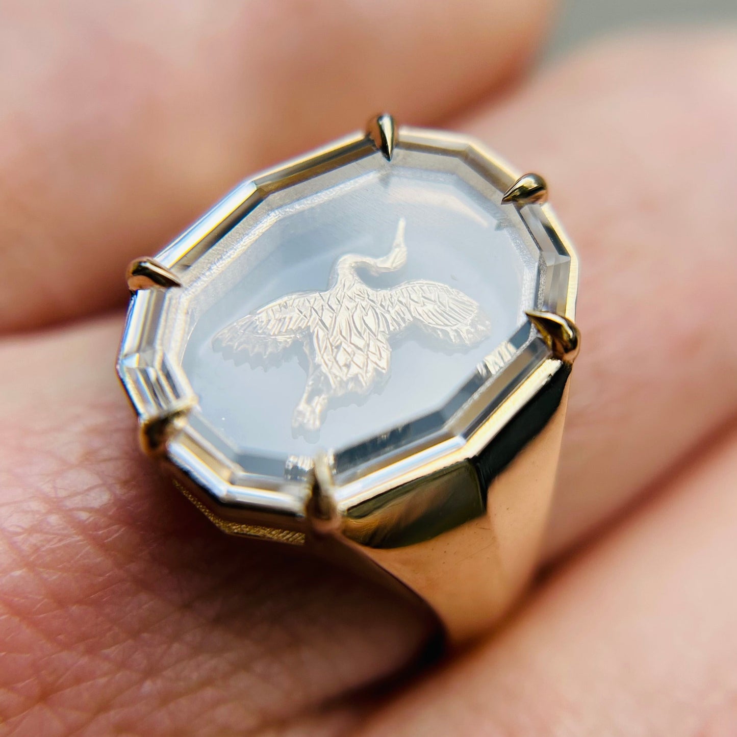 A gold ring with an octagonal bezel featuring a detailed engraving of a Japanese crane bird in flight on a translucent stone, shown worn on a finger.