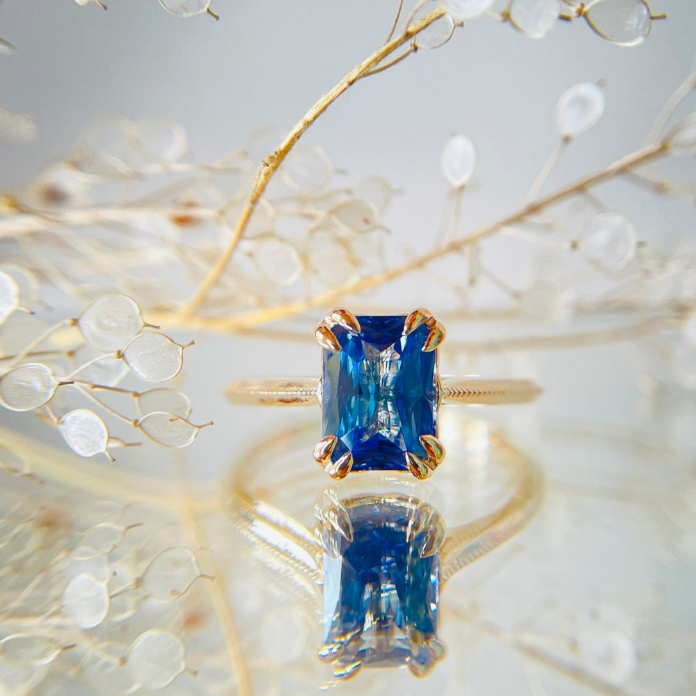 A yellow gold engagement ring with a radiant blue sapphire is elegantly displayed. The background features delicate, transparent leaves, adding a refined, serene touch.