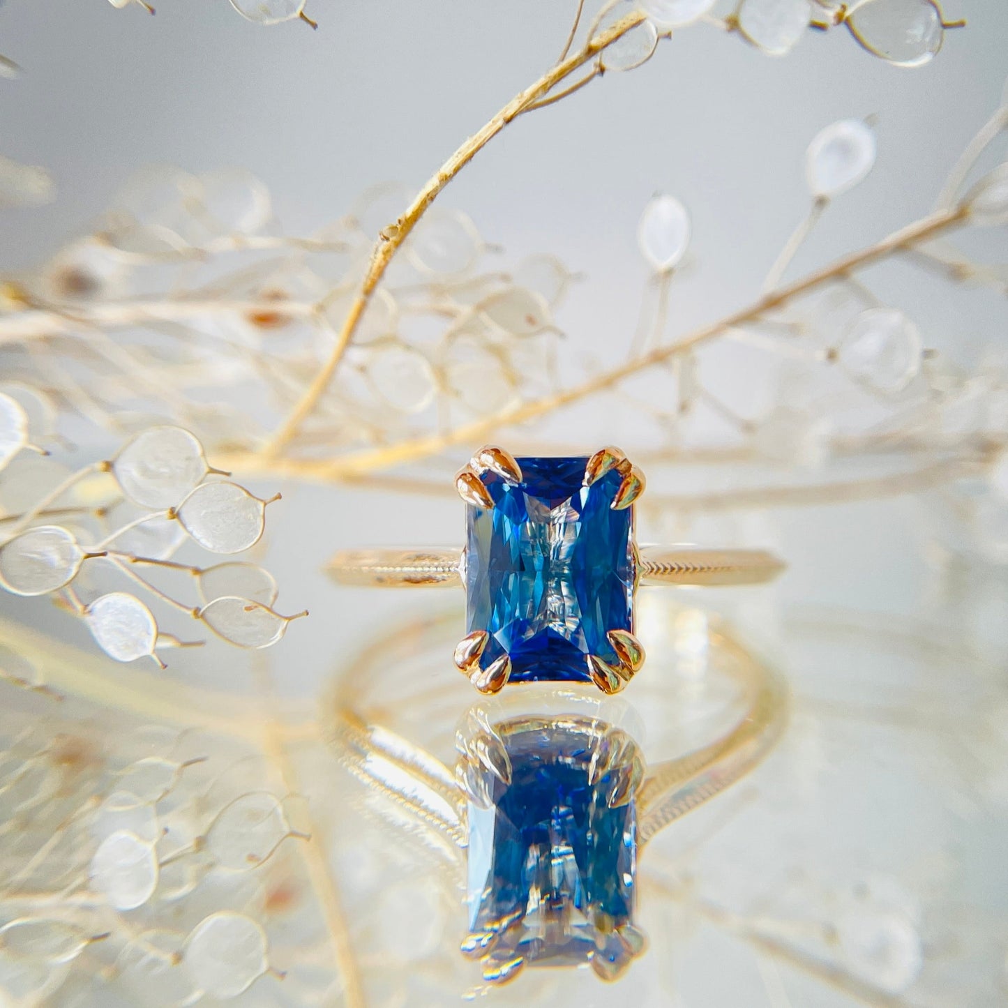 A yellow gold engagement ring with a radiant blue sapphire is elegantly displayed. The background features delicate, transparent leaves, adding a refined, serene touch.