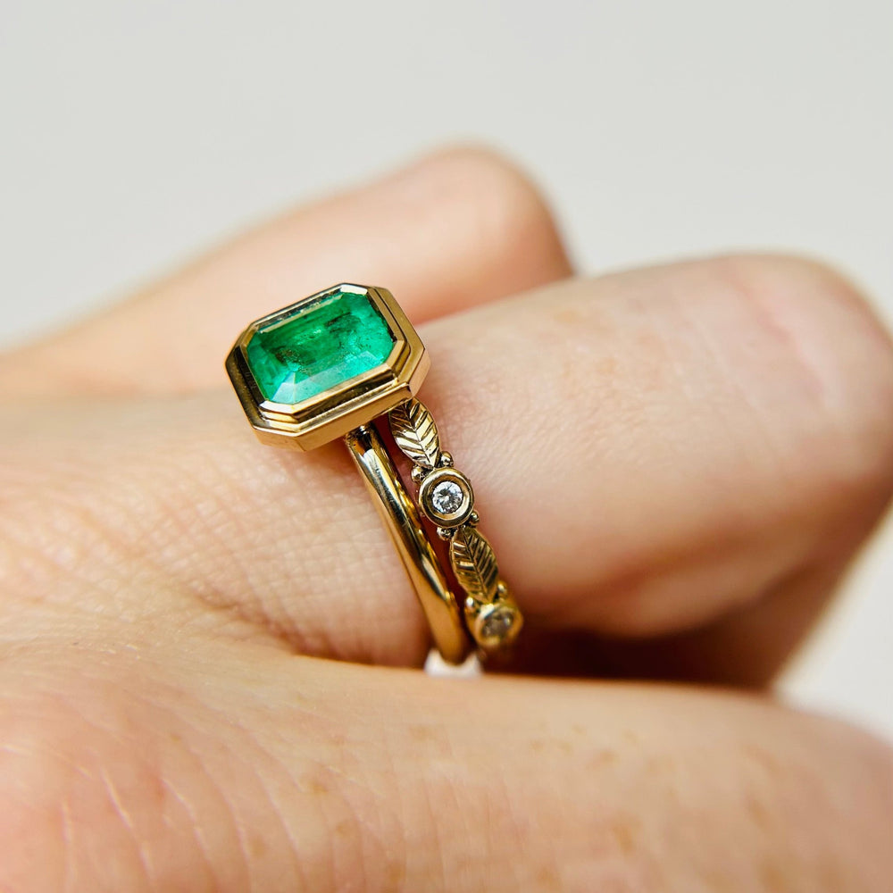 
                  
                    An emerald solitaire engagement ring stacked with a gold wedding band, shown on a finger.
                  
                