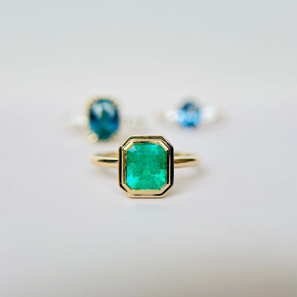 
                  
                    An emerald solitaire bezel set engagement ring is in the foreground and blurred in the background are two blue engagement rings in soft focus. Resting on a bright surface.
                  
                