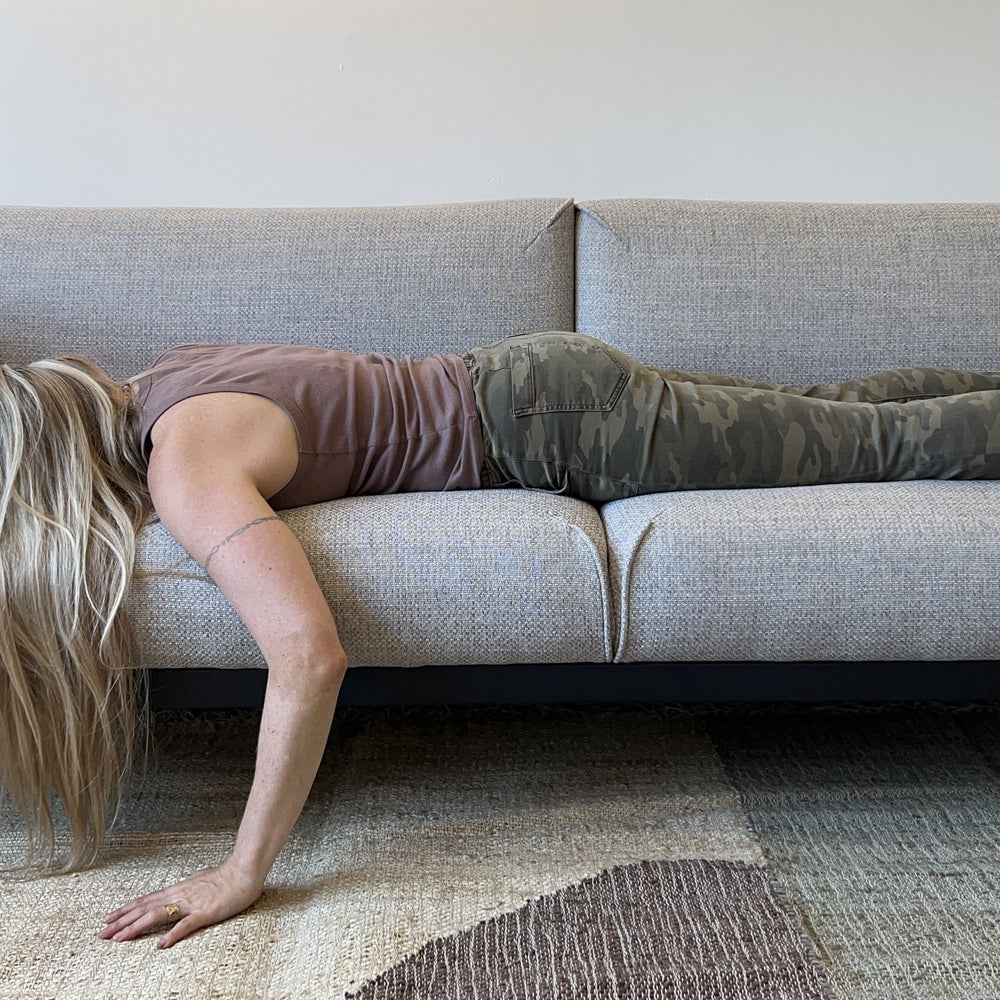 A woman in camo pants and a tank top lies face down on a gray sofa, arm hanging over the side. The tone of the image is humorous and lighthearted, her posture suggests comfort, being at home and relaxation.