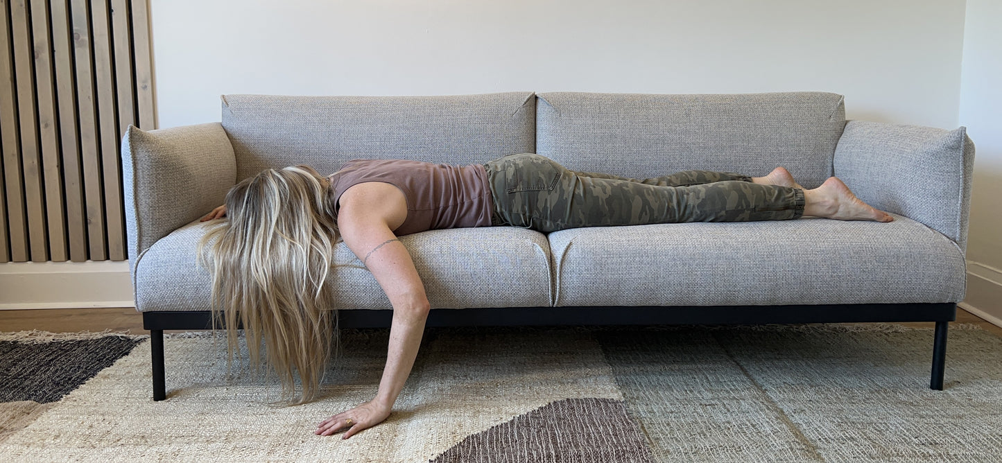A woman in camo pants and a tank top lies face down on a gray sofa, arm hanging over the side. The tone of the image is humorous and lighthearted, her posture suggests comfort, being at home and relaxation.
