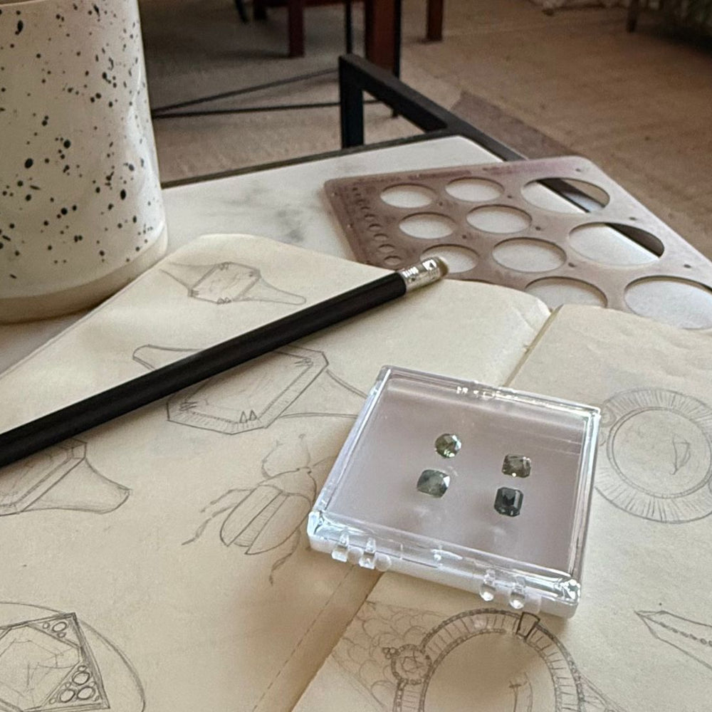 A cozy workspace with a speckled mug, pencil, and jewellery sketches on a notebook. Nearby, a case displays four small gemstones, conveying creativity and inspiration.