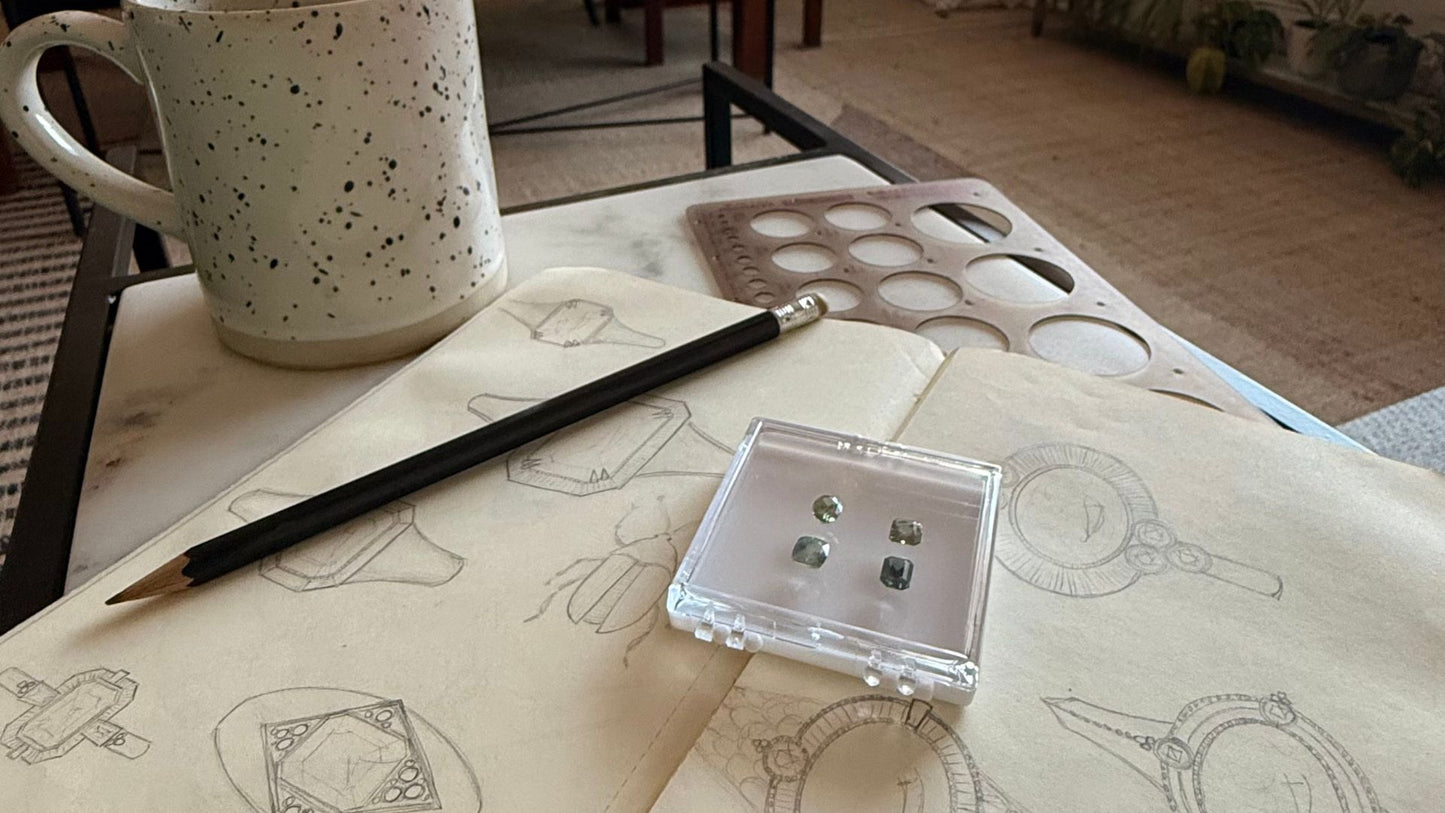 A cozy workspace with a speckled mug, pencil, and jewellery sketches on a notebook. Nearby, a case displays four small gemstones, conveying creativity and inspiration.