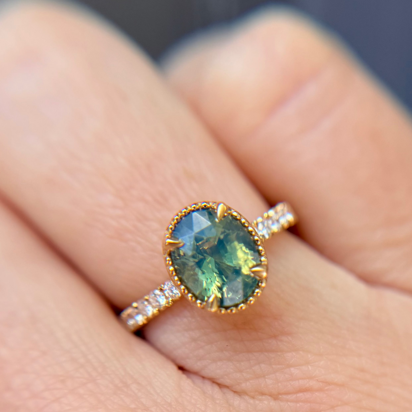 Yellow Gold ring with a green and teal sapphire centre stone with diamond accents along the band, shown worn on a person's finger