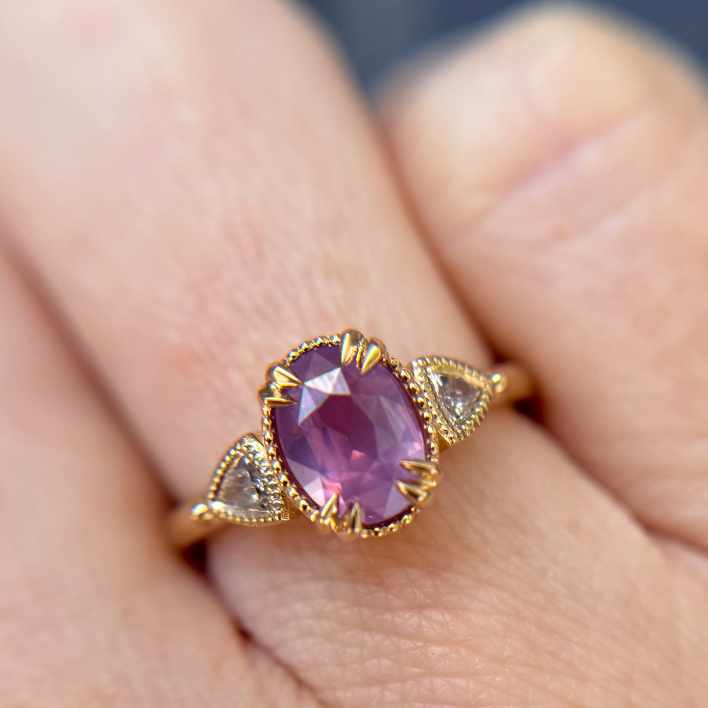 Yellow Gold 3-stone engagement ring with a purple pink sapphire gemstone worn on a finger. Featuring diamond accents either side of the oval center stone.