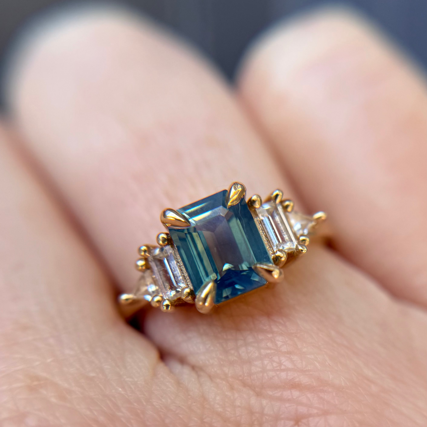 Yellow Gold engagement ring with a teal sapphire set against white accent stones worn on a finger.