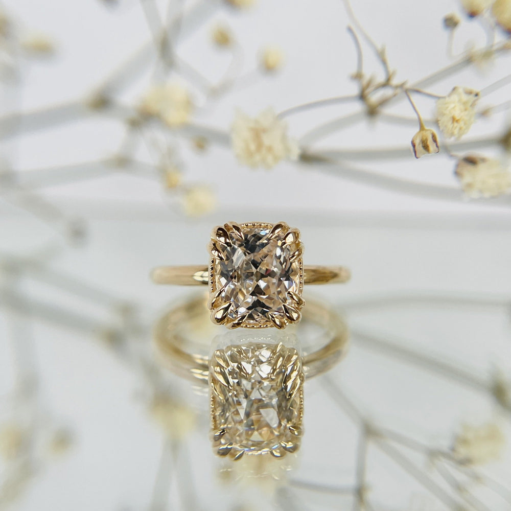 A gold ring with a large, cushion cut diamond is reflected on a glossy surface. Delicate dried flowers in the background create an elegant, romantic tone.