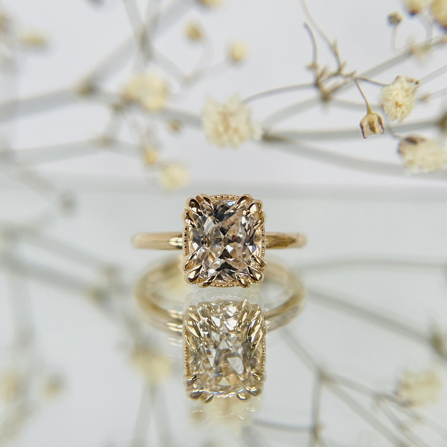A gold ring with a large, cushion cut diamond is reflected on a glossy surface. Delicate dried flowers in the background create an elegant, romantic tone.