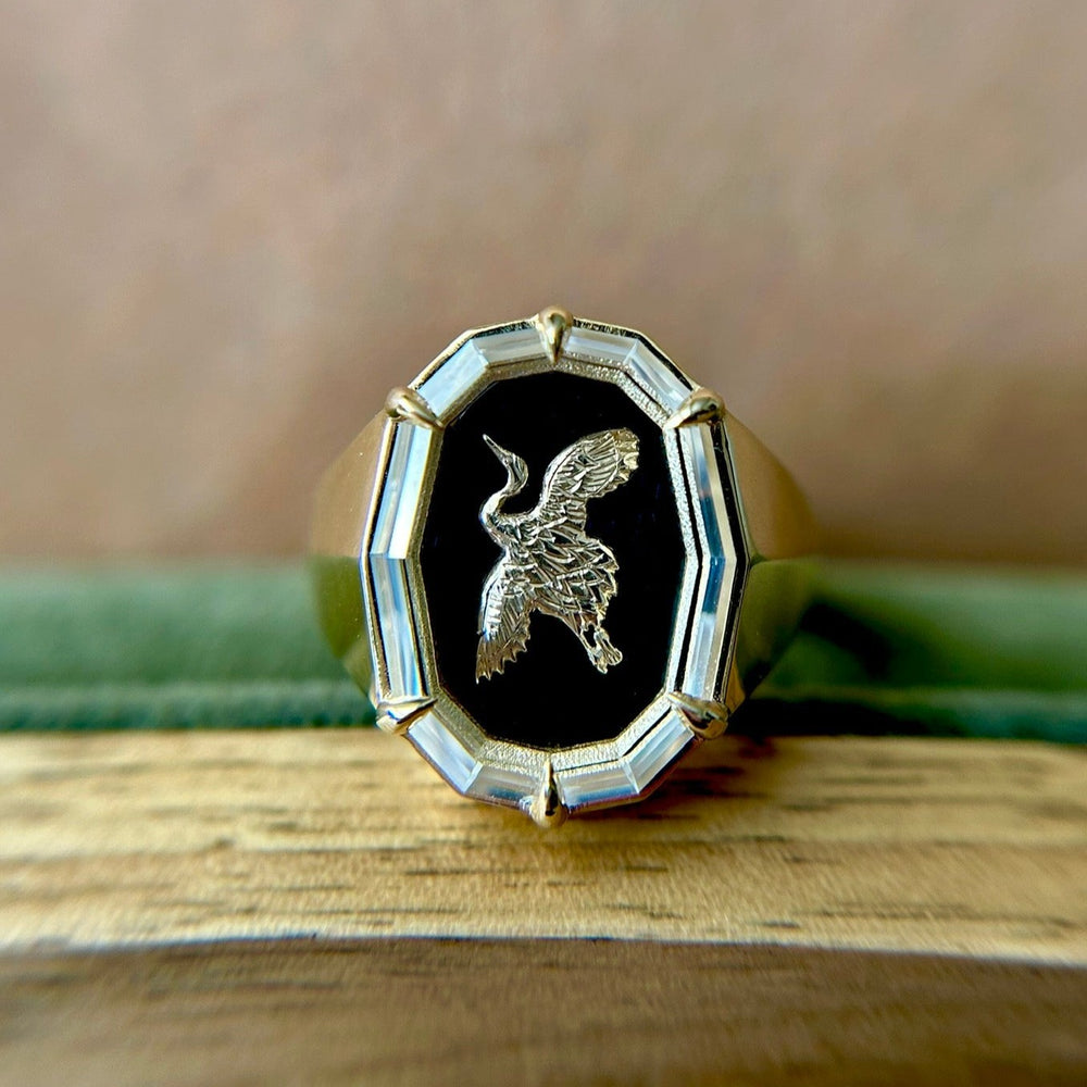 Gold signet ring with an engraved bird in flight on a black onyx background, framed by a geometric border. The ring rests on a wooden surface, conveying elegance.