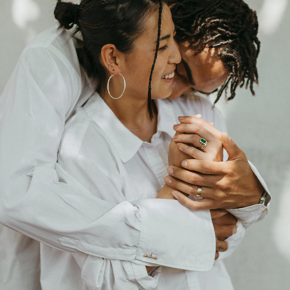 A couple embraces warmly, smiling and holding hands. Both wear white shirts and gold engagement rings. His is a simple matte finish gold wedding ring and hers is a solitaire engagement ring with a large emerald. The scene conveys love and comfort with soft, dappled sunlight.