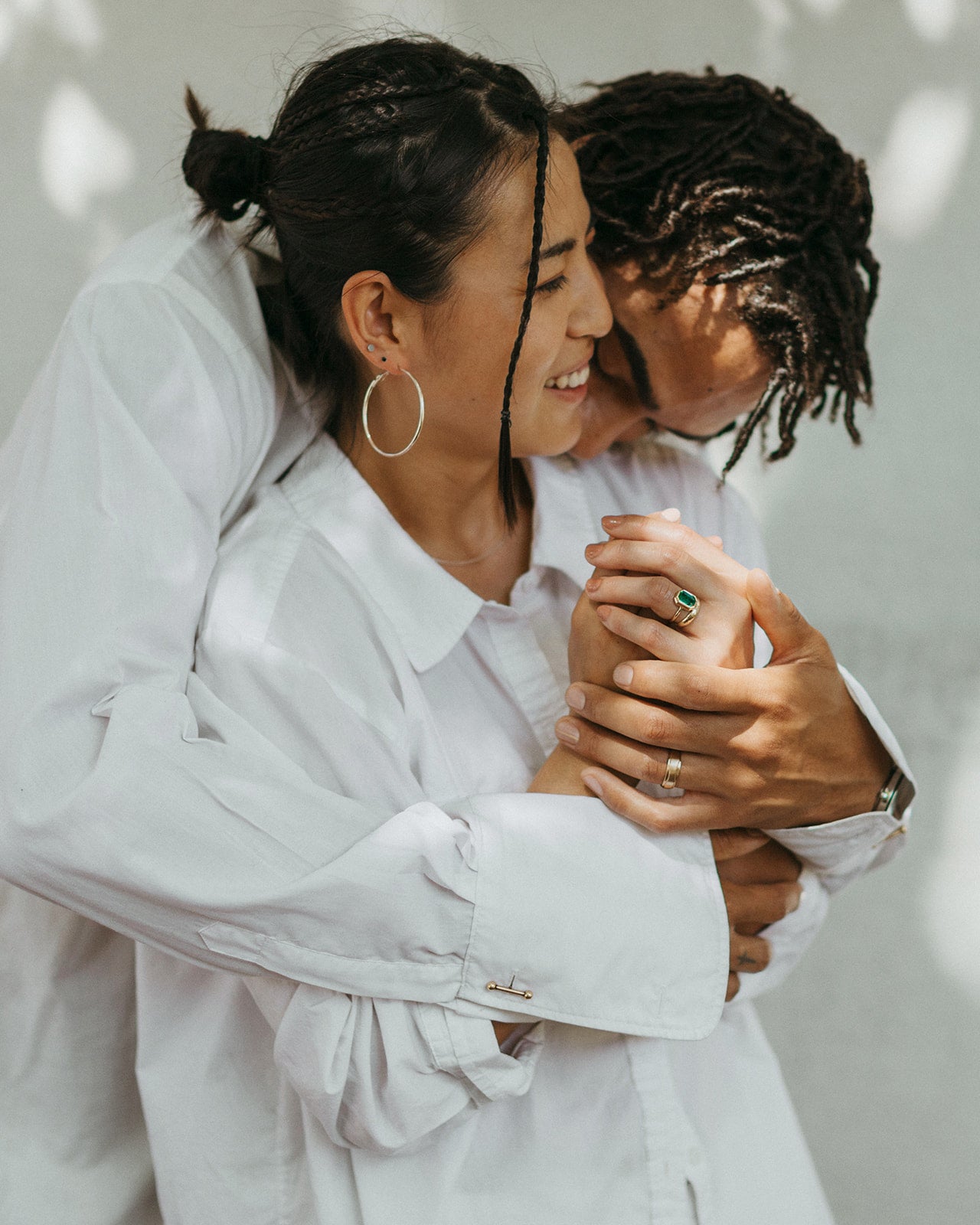 A couple embraces warmly, smiling and holding hands. Both wear white shirts and gold engagement rings. His is a simple matte finish gold wedding ring and hers is a solitaire engagement ring with a large emerald. The scene conveys love and comfort with soft, dappled sunlight.