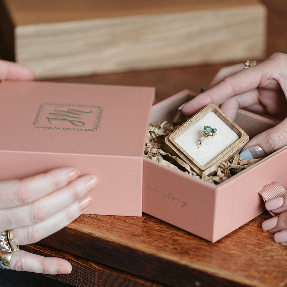 Two hands hold a pink box with a monogrammed lid with the Jilian Maddin Jewellery logo, revealing a ring with a green sapphire inside on a wooden table. The scene conveys elegance and anticipation.