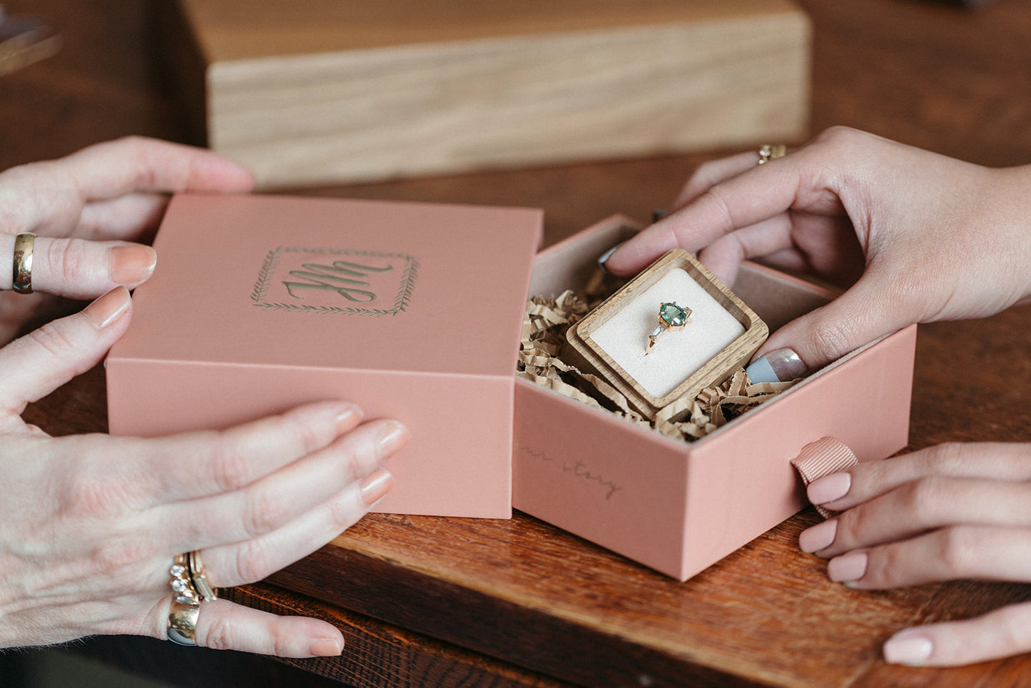 Two hands hold a pink box with a monogrammed lid with the Jilian Maddin Jewellery logo, revealing a ring with a green sapphire inside on a wooden table. The scene conveys elegance and anticipation.