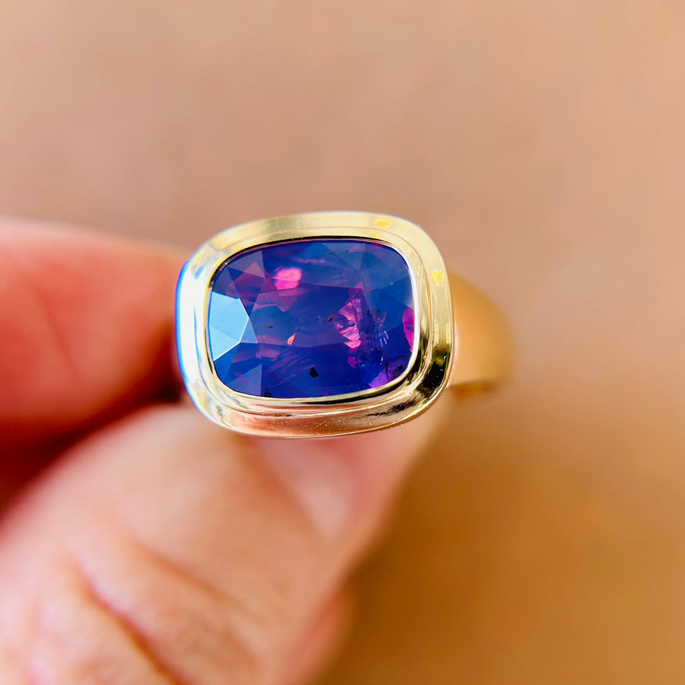 A gold bezel set ring with a vibrant, faceted purple sapphire is held delicately between fingers. The gem glows, conveying elegance and luxury against a blurred background.