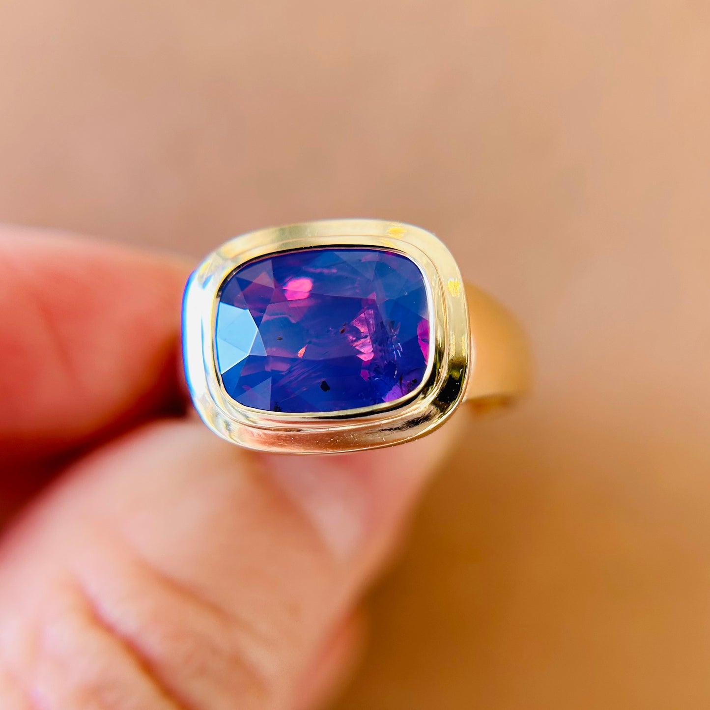 A gold bezel set ring with a vibrant, faceted purple sapphire is held delicately between fingers. The gem glows, conveying elegance and luxury against a blurred background.
