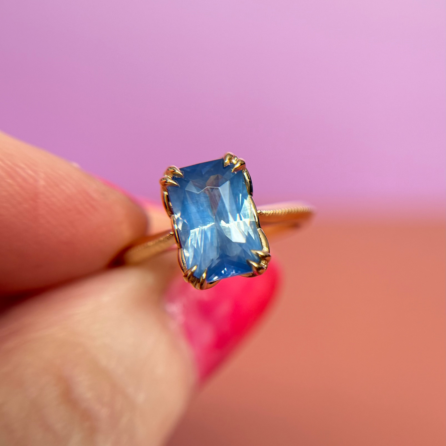Yellow Gold solitaire engagement ring with a blue sapphire centre stone held between fingers against a pink background