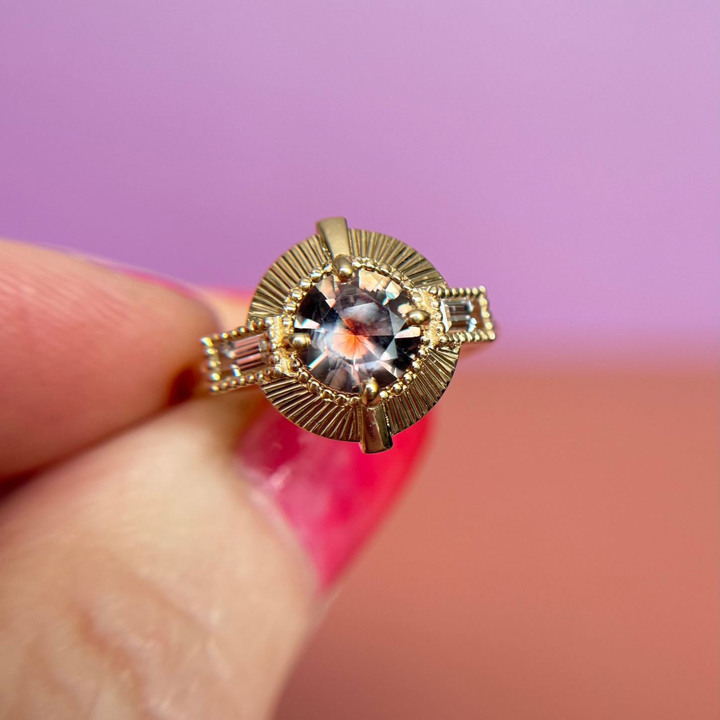 Gold ring with a round pink sapphire gemstone held between fingers against a pink background