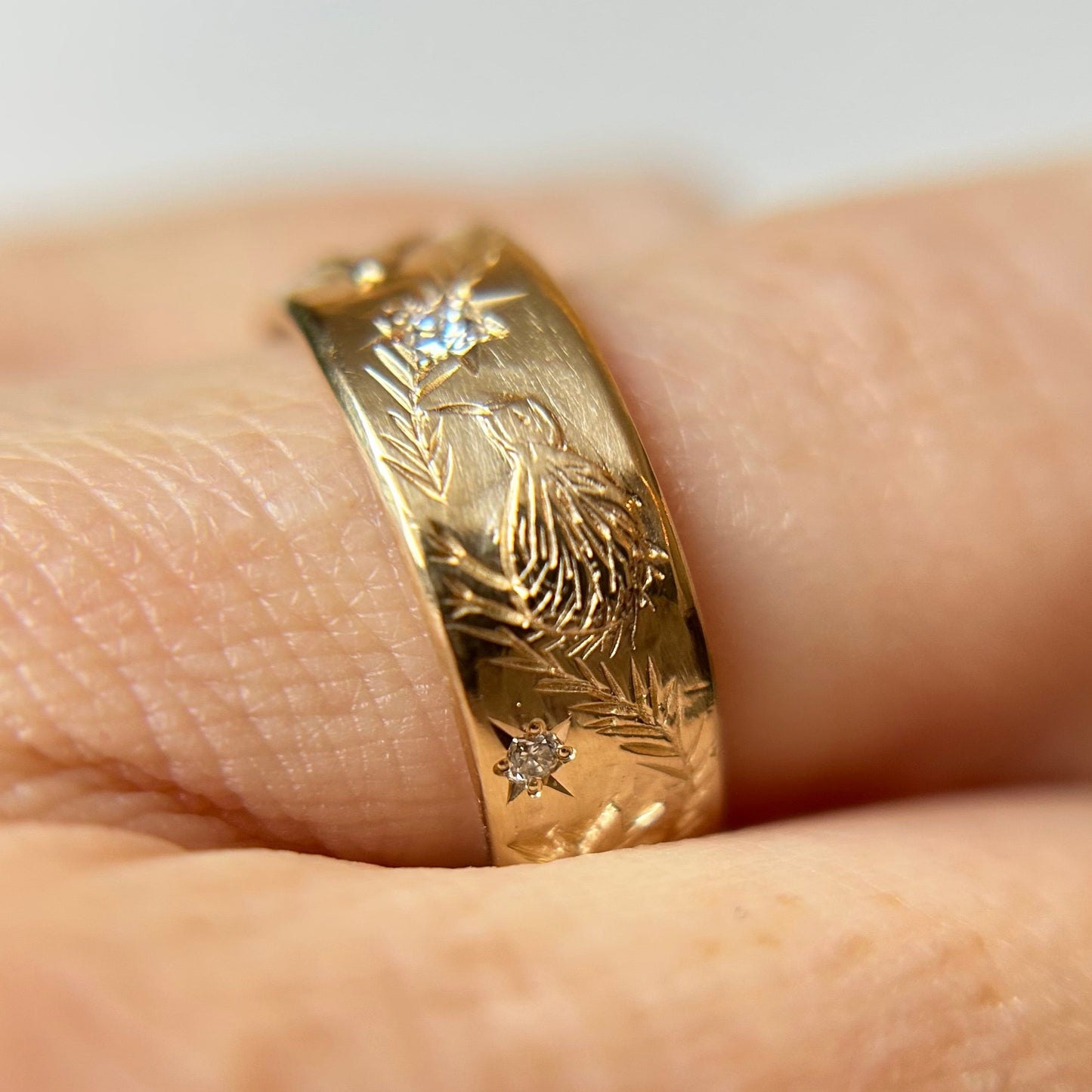 Close-up of a 14K gold ring on a finger, featuring intricate engraved bird and leaf patterns and small sparkling diamonds. The design conveys elegance and unique handmade craftsmanship.