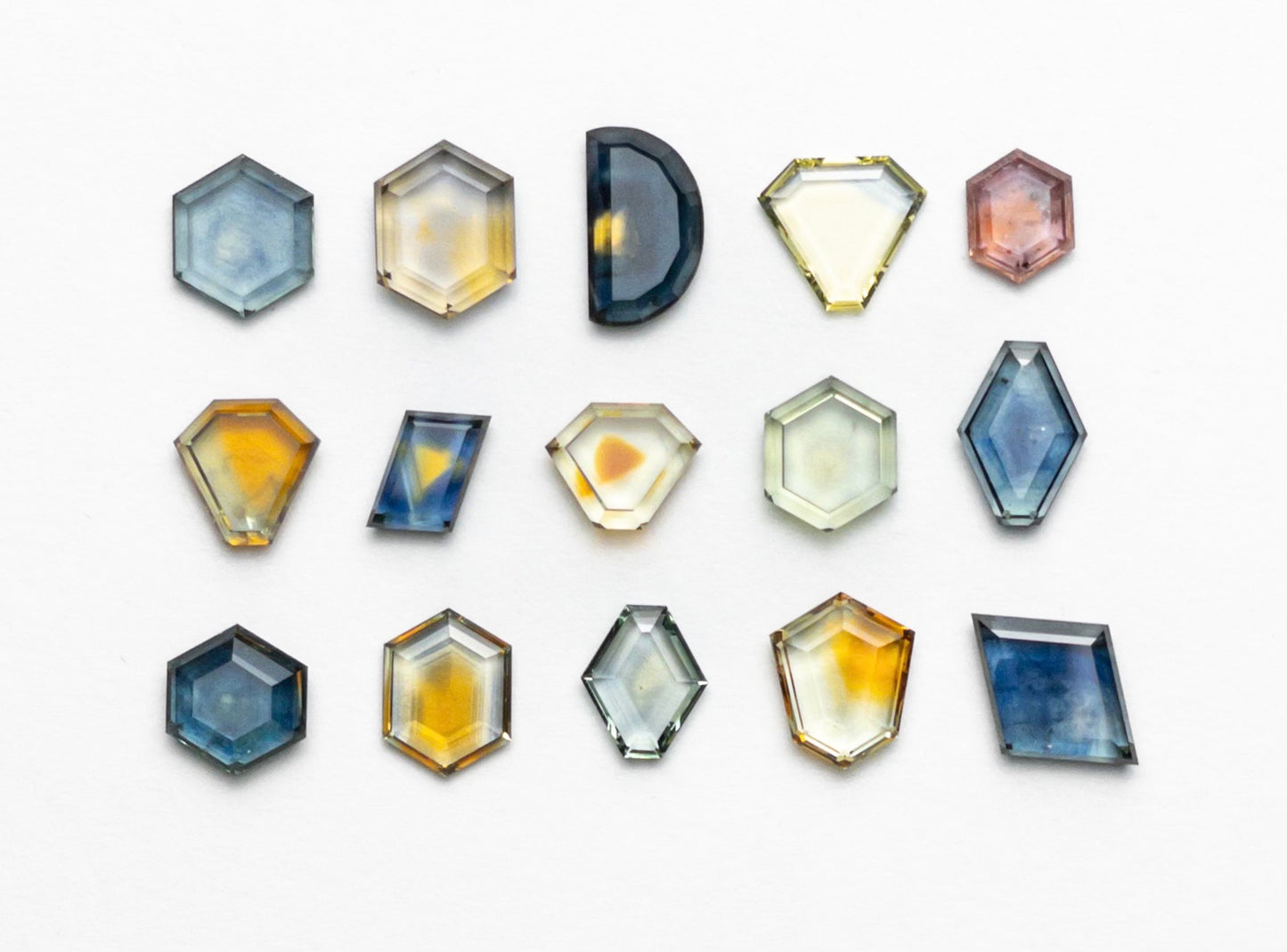 A collection of colorful, multi-shaped gemstones is arranged neatly on a white background. The stones are in hexagonal, octagonal, and half-circle shapes, featuring hues of blue, yellow, pink, and orange, creating a vibrant visual display.
