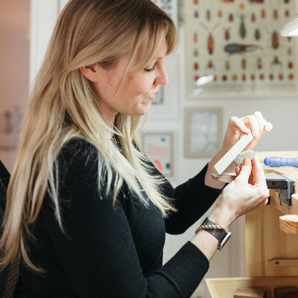 Jilian Maddin sits at her jewellery bench in her well-lit Toronto studio filing metal, expressing concentration. Background shows insect illustrations and an inviting atmosphere.