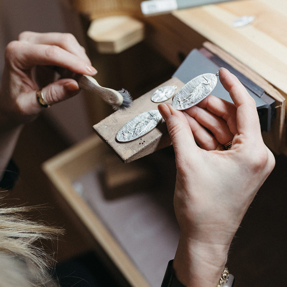 Jilian Maddin is holding a wooden block with engraved silver pendants, using a small brush for detailing. Jewelry crafting tools are visible in a workshop setting.