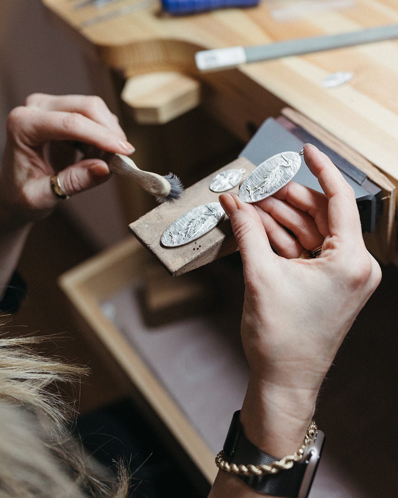 Jilian Maddin is holding a wooden block with engraved silver pendants, using a small brush for detailing. Jewelry crafting tools are visible in a workshop setting.