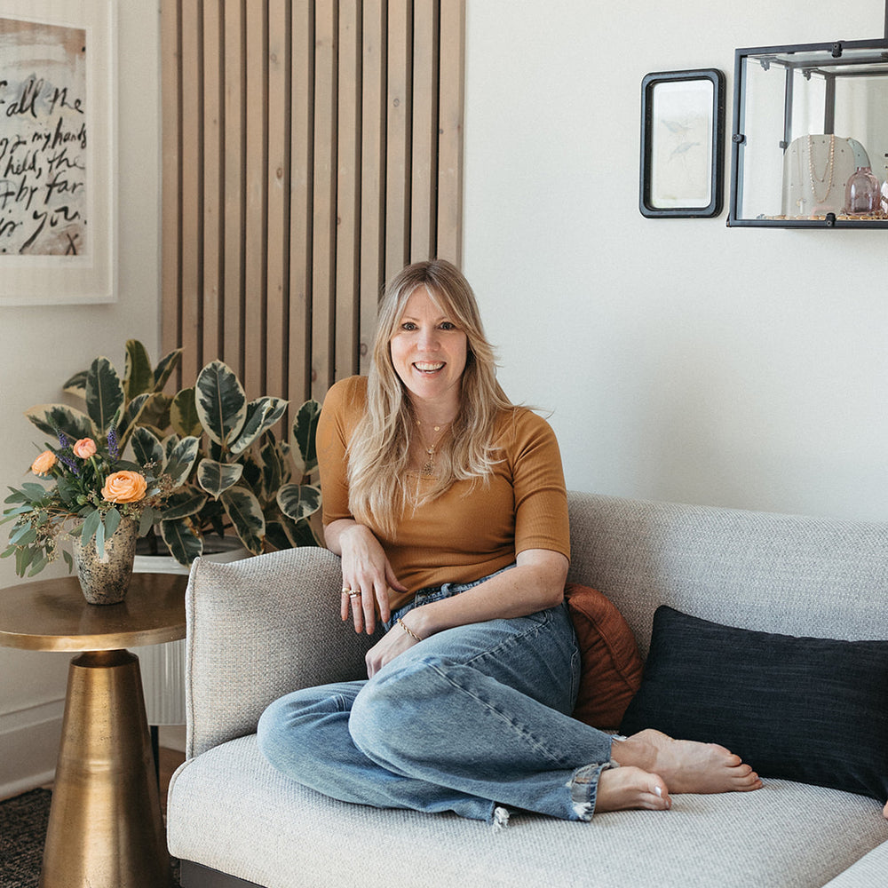 Jilian Maddin sits casually on a beige couch in her Toronto Jewellery studio, smiling warmly. The room has a modern, cozy feel with plants, art, jewellery in glass display cases on the wall and a side table with peachy orange flowers.